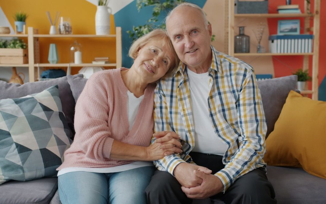 Elderly couple smiling and holding hands on couch.